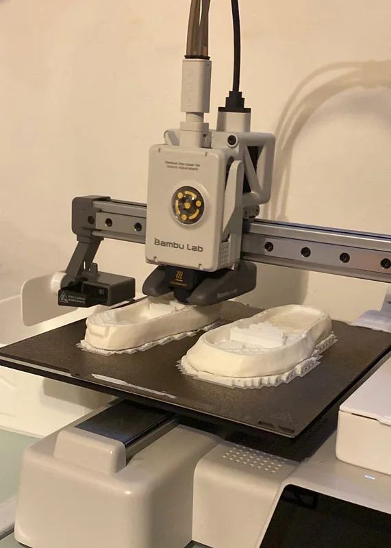 A 3D printer from Bambu Lab is creating two shoe molds on a build plate, situated in a well-lit room.